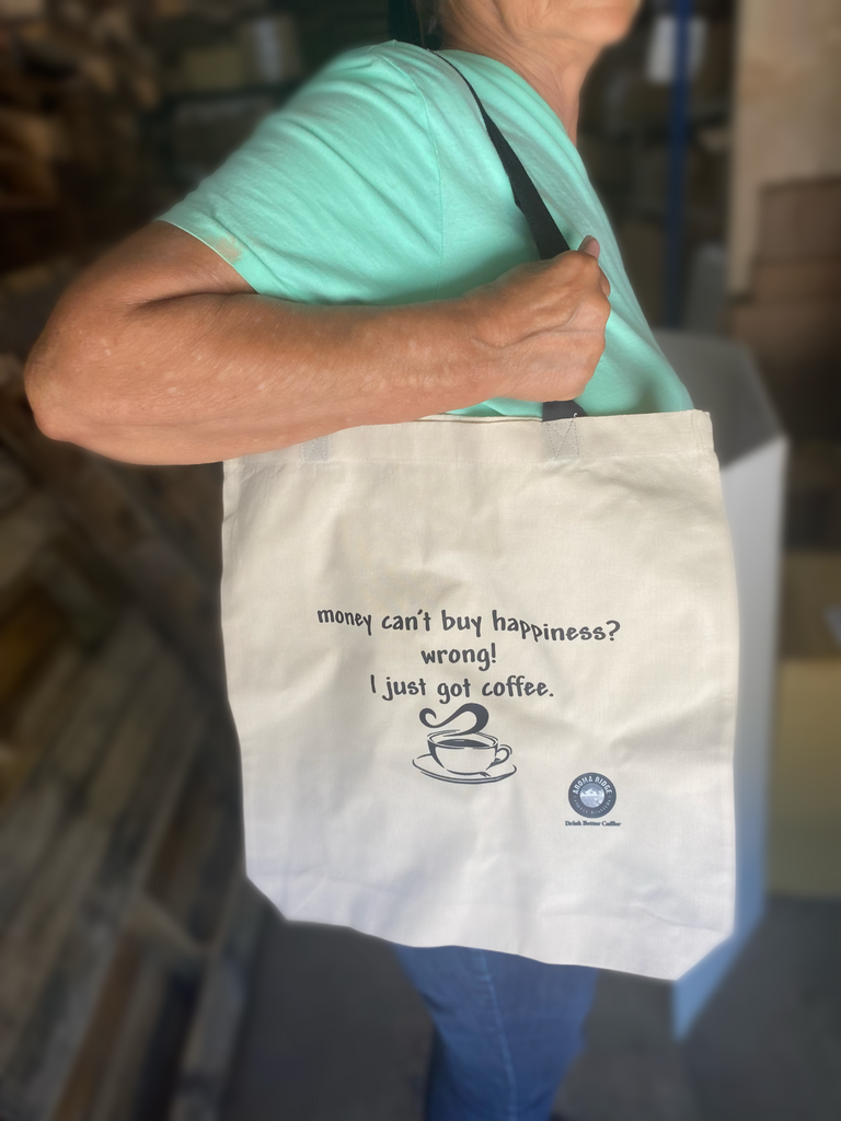 A person holding a beige cotton tote bag with a printed message 'Money can't buy happiness? I just got coffee'.